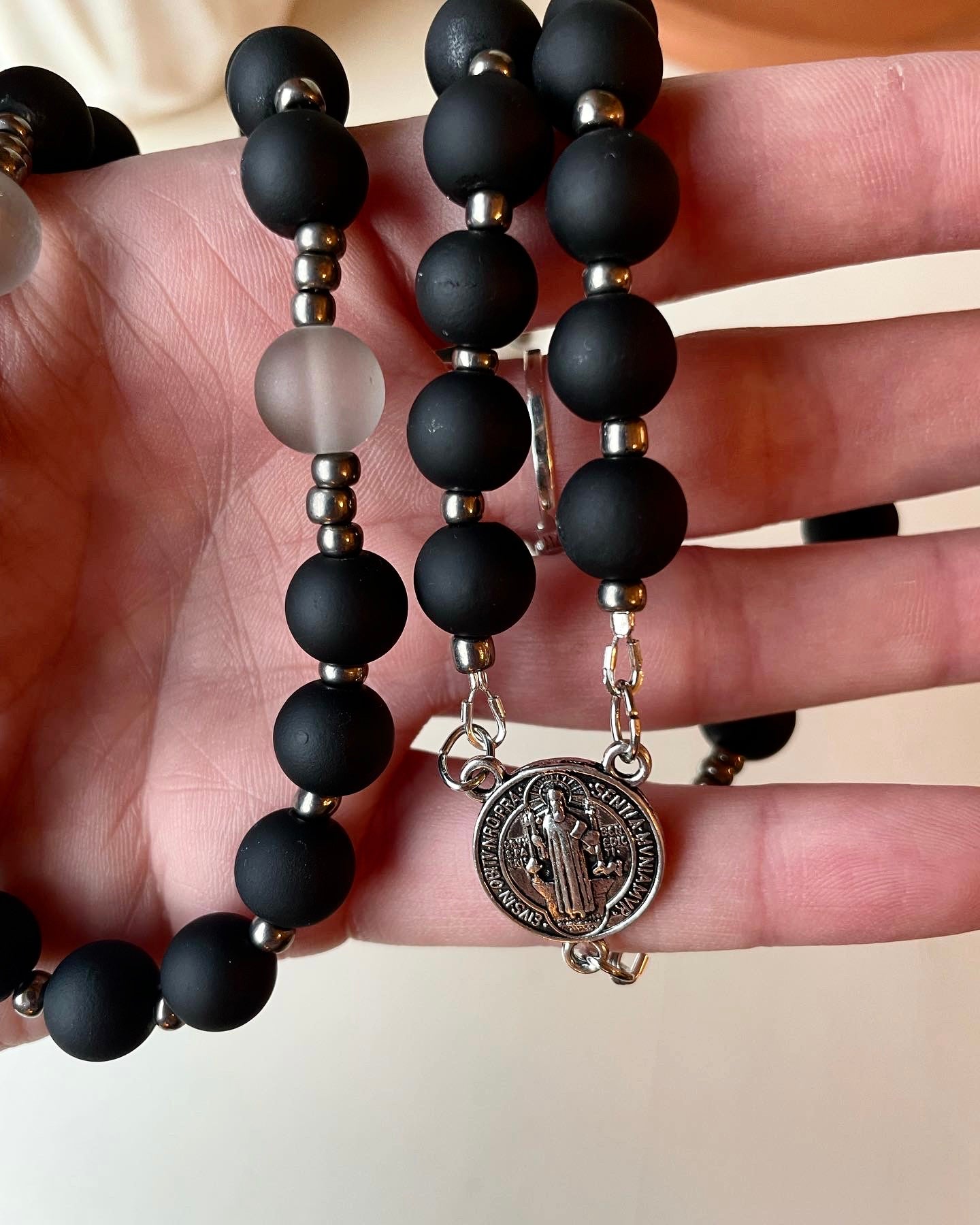 This heavyweight rosary is comprised of a matte black glass bead with a slightly rubberized texture. A light grey glass bead is added for the Our Father prayers, and is completed with silver gunmetal seed beads as spacers for a seamless experience.  This set has a silver pewter St. Benedict Medal center and a Pardon Crucifix to finish it off.